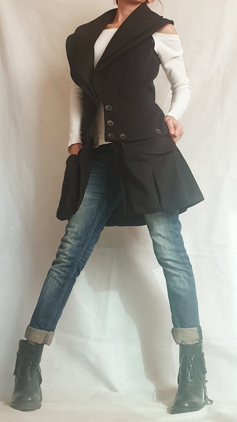 May include: A black sleeveless vest with a large collar and a pleated skirt. The vest has a double breasted closure with buttons. The person is wearing blue jeans and black boots.