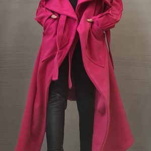 K&ouml;nnte beinhalten: Ein langer, leuchtend pinker Mantel mit einem breiten, drapierten Kragen, langen &Auml;rmeln und einem Bindeg&uuml;rtel in der Taille. Der Mantel wird &uuml;ber einem schwarzen Oberteil, einer schwarzen Hose und schwarzen Schn&uuml;rstiefeln getragen.