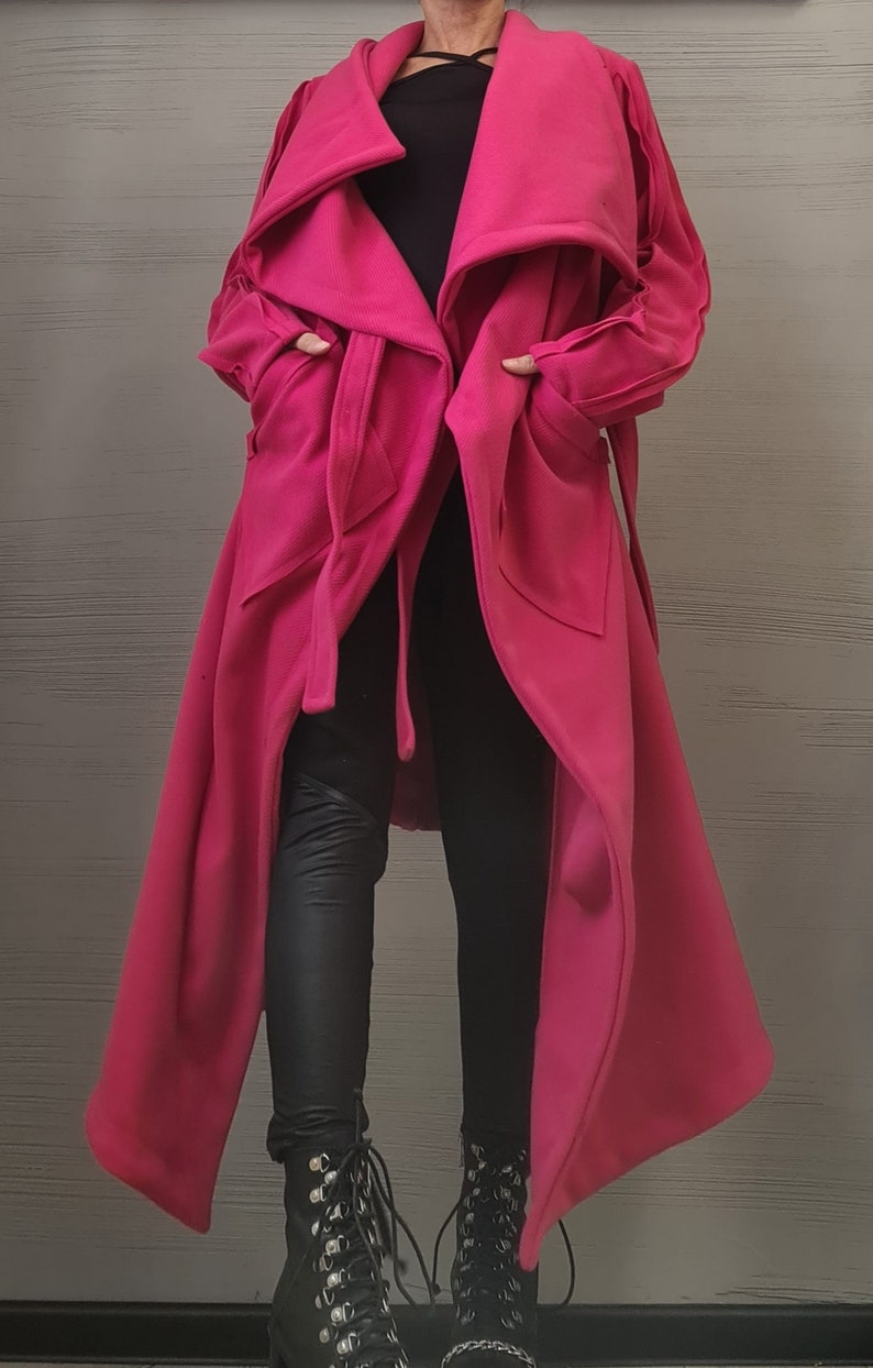 K&ouml;nnte beinhalten: Ein langer, leuchtend pinker Mantel mit einem breiten, drapierten Kragen, langen &Auml;rmeln und einem Bindeg&uuml;rtel in der Taille. Der Mantel wird &uuml;ber einem schwarzen Oberteil, einer schwarzen Hose und schwarzen Schn&uuml;rstiefeln getragen.