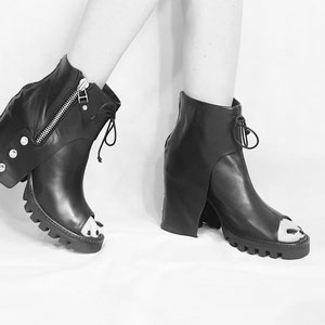 May include: Black leather peep-toe ankle boots with a chunky heel and a zipper detail on the side. The boots have a unique design with a flap that extends from the ankle to the heel.