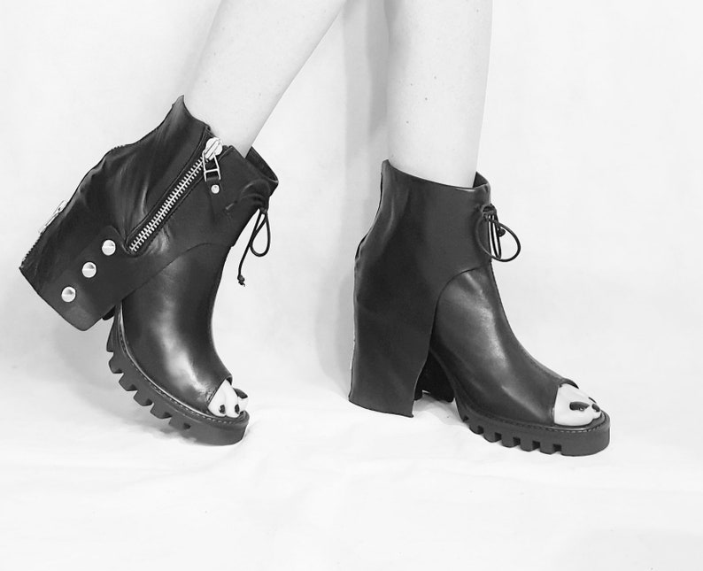May include: Black leather peep-toe ankle boots with a chunky heel and a zipper detail on the side. The boots have a unique design with a flap that extends from the ankle to the heel.