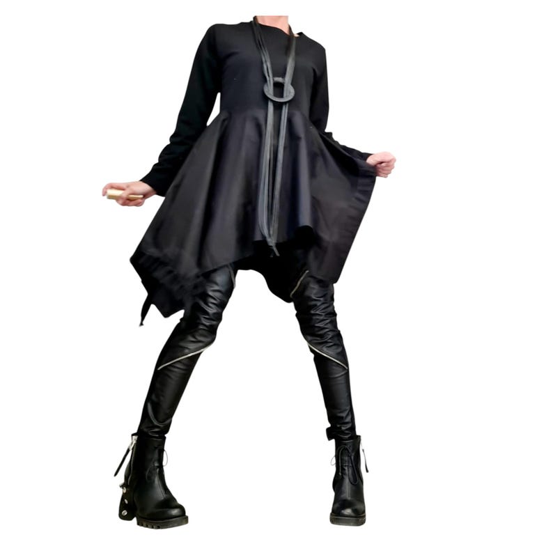 May include: A black asymmetrical dress with long sleeves and a flared skirt. The outfit includes black leather pants with silver zippers and black ankle boots. A black necklace with a large circular pendant completes the look.
