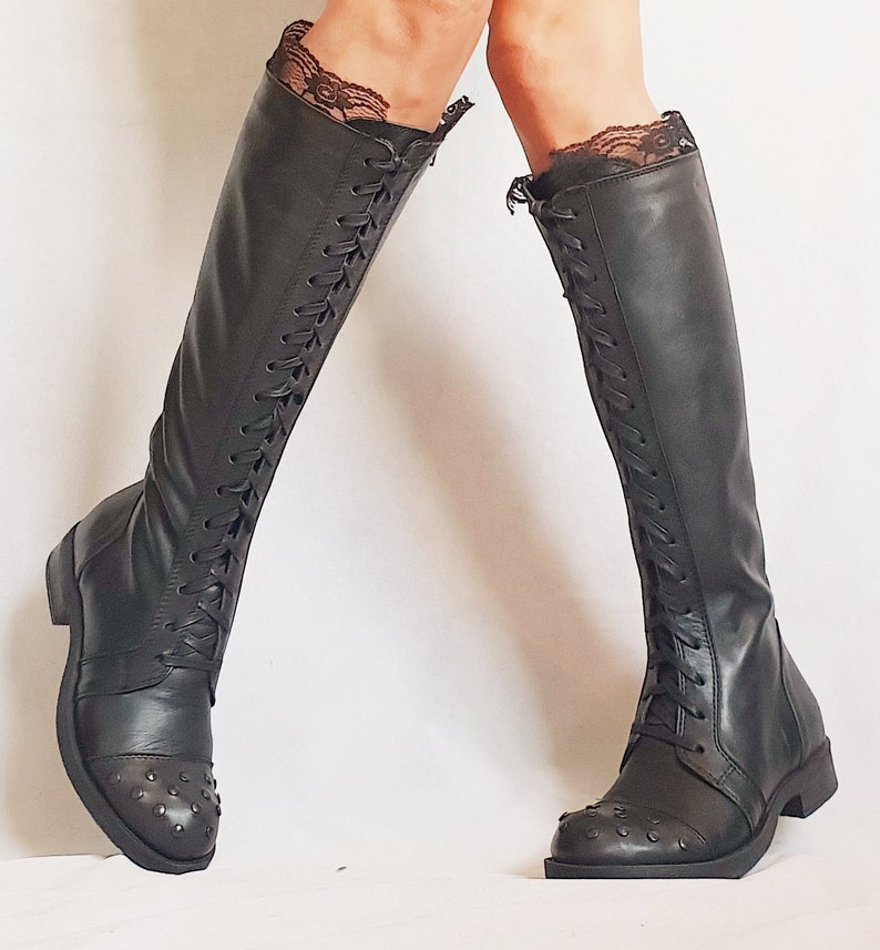 May include: Black knee-high boots with lace trim at the top. The boots have a lace-up design on the front and small studs on the toe. The boots have a flat sole.