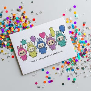 May include: A white greeting card with a colorful illustration of six cartoon characters in bunny costumes, each holding a balloon. The card reads "HAVE A FABU-LABUBU-US BIRTHDAY!" and is surrounded by colorful confetti.