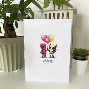 Può includere: Biglietto di auguri bianco con illustrazioni di Deadpool e Wolverine che tengono dei palloncini. Il testo "Oh. My F**k! It's your Birthday!" è stampato sotto. Il biglietto è esposto su una superficie bianca.