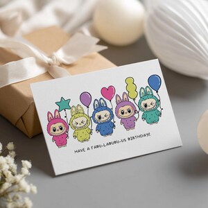 May include: A white greeting card with a cartoon illustration of five bunny-like characters in various colors, each holding a balloon. The card reads "HAVE A FABU-LABUBU-US BIRTHDAY!" in black text.