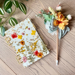 Puede incluir: Un cuaderno de espiral dorado con una cubierta transparente que presenta flores prensadas y pan de oro. El cuaderno está sobre una superficie de madera junto a un bolígrafo decorativo con un lazo azul y un ramo de flores.