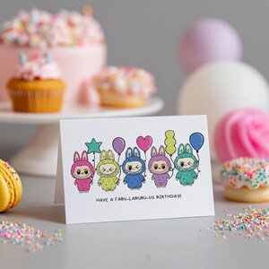 May include: A white greeting card with a cartoon illustration of six characters in bunny costumes holding balloons. The characters are in various colors, including pink, yellow, blue, purple, and green. The text "HAVE A FABU-LABUBU-US BIRTHDAY!" is printed below the characters. The background includes a cake, cupcakes, and macarons.
