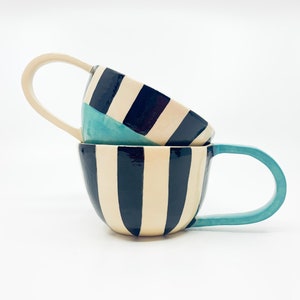 May include: Two ceramic mugs with black and white striped patterns. The top mug has a turquoise blue accent. The mugs are stacked on top of each other.