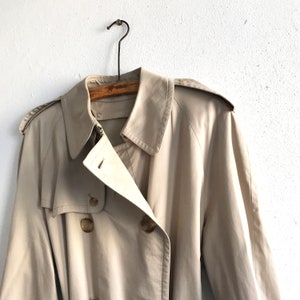 K&ouml;nnte beinhalten: Ein beiger Trenchcoat mit doppelreihigem Verschluss und G&uuml;rtel. Der Mantel hat ein klassisches Design und ist aus leichtem Stoff gefertigt.