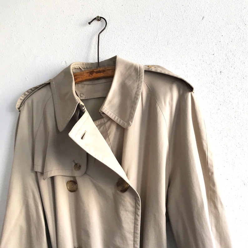 K&ouml;nnte beinhalten: Ein beiger Trenchcoat mit doppelreihigem Verschluss und G&uuml;rtel. Der Mantel hat ein klassisches Design und ist aus leichtem Stoff gefertigt.