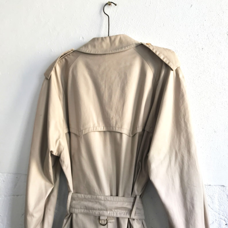 K&ouml;nnte beinhalten: Ein beiger Trenchcoat mit G&uuml;rtel und einem einzigen Knopf auf dem R&uuml;cken. Der Mantel h&auml;ngt an einem Kleiderb&uuml;gel an einer wei&szlig;en Wand.