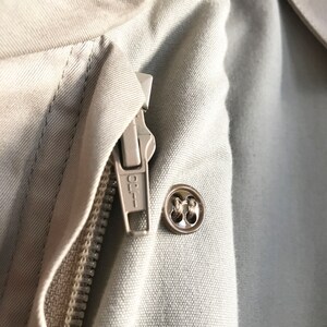 K&ouml;nnte beinhalten: Nahaufnahme einer beige Jacke mit Rei&szlig;verschluss und Knopf. Der Rei&szlig;verschluss hat den Schriftzug "OPT" aufgedruckt.