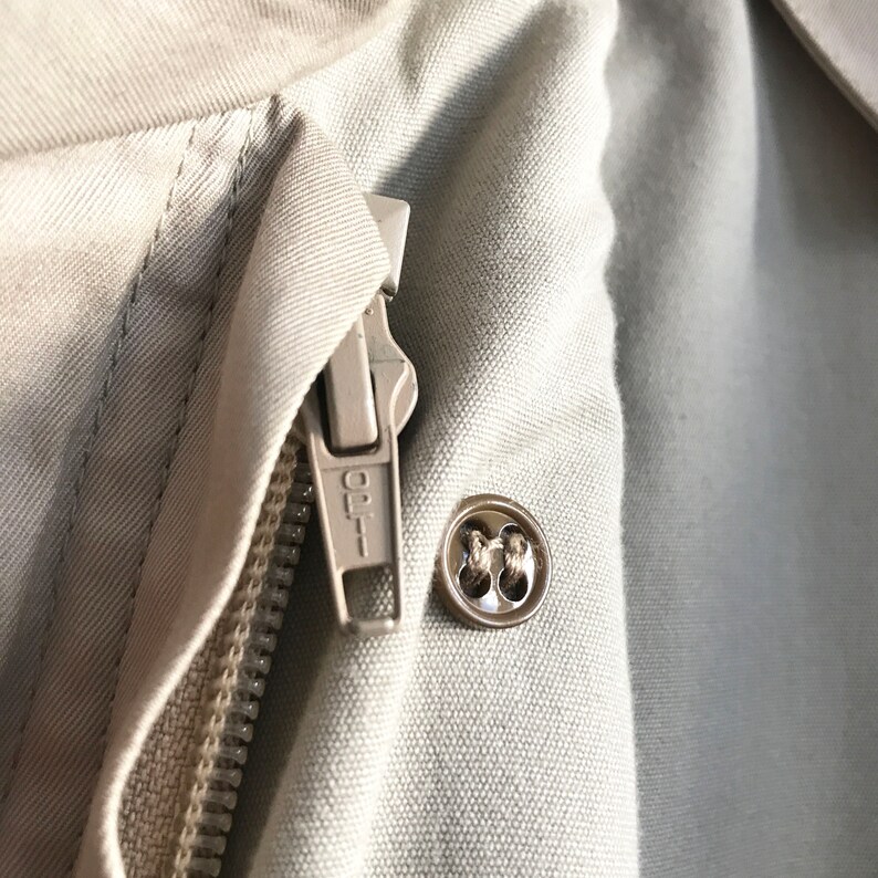 K&ouml;nnte beinhalten: Nahaufnahme einer beige Jacke mit Rei&szlig;verschluss und Knopf. Der Rei&szlig;verschluss hat den Schriftzug "OPT" aufgedruckt.