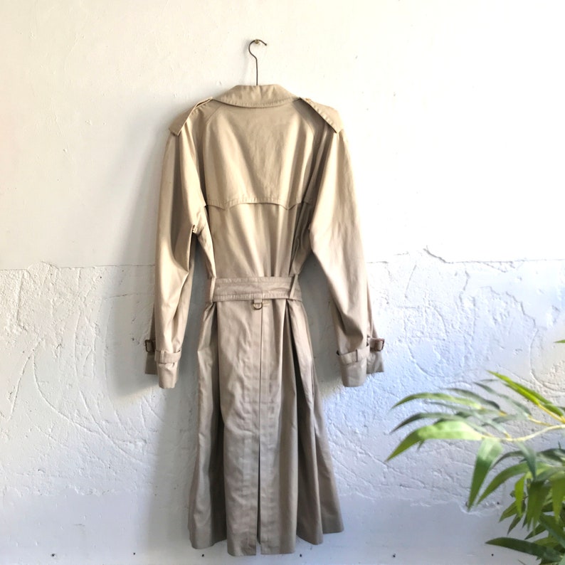 K&ouml;nnte beinhalten: Ein beiger Trenchcoat mit G&uuml;rtel und einem Kleiderb&uuml;gel. Der Mantel h&auml;ngt an einer wei&szlig;en Wand.