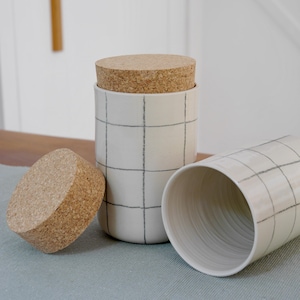 May include: A set of three ceramic canisters with cork lids. The canisters are off-white with a hand-drawn black grid pattern. One canister is upright with a lid, another is on its side, and the third lid is separate.