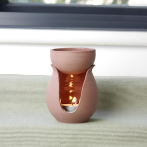 May include: A terracotta essential oil diffuser with a candle inside. The diffuser has a rounded base with a flared opening and a separate top bowl. The candle flame is visible through the opening, creating a warm glow.