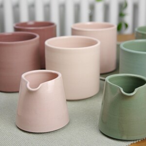 May include: A collection of ceramic pitchers and cylindrical mugs in various pastel colors, including pink, beige, and green. The pitchers have a spout for pouring. The mugs are simple and modern.