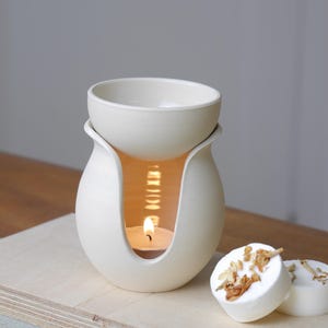 May include: A cream ceramic essential oil diffuser with a candle inside. The diffuser has a round bowl on top and a curved design. Two white wax melts with dried flowers are next to the diffuser. The candle flame is visible through the opening.