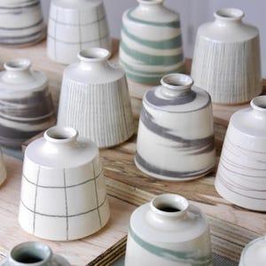 May include: A collection of small, white ceramic vases with various gray and green painted designs. Some vases have striped, swirled, or grid patterns. The vases are arranged on a light brown wooden surface.