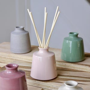 May include: A collection of ceramic reed diffuser bottles in various colors, including pink, gray, and green. The pink bottle in the center holds several light-colored diffuser reeds. The bottles are arranged on a wooden surface.