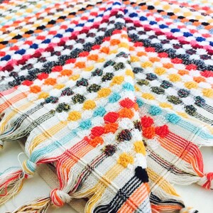 May include: A colorful, patterned textile with a geometric design. The fabric features a grid of small, raised dots in various colors, including red, orange, yellow, blue, and white. The edges are finished with knotted tassels in coordinating colors. This item is suitable for home decor.