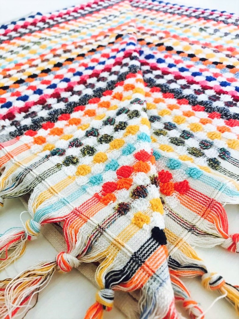 May include: A colorful, patterned textile with a geometric design. The fabric features a grid of small, raised dots in various colors, including red, orange, yellow, blue, and white. The edges are finished with knotted tassels in coordinating colors. This item is suitable for home decor.