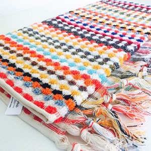 May include: A folded, colorful woven towel with a textured, dotted pattern. The towel features a variety of colors including red, orange, yellow, blue, and white. The edges are finished with a fringe of multicolored threads.
