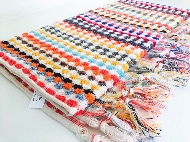 May include: A folded, colorful woven towel with a textured, dotted pattern. The towel features a variety of colors including red, orange, yellow, blue, and white. The edges are finished with a fringe of multicolored threads.