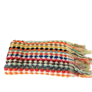May include: A colorful, patterned blanket with a textured, dotted design. The blanket features a mix of colors including orange, blue, pink, and white. The edges are finished with fringe.