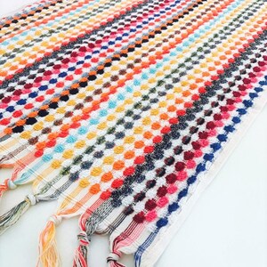 May include: A colorful, striped towel with a textured, dotted pattern. The towel features a variety of colors, including orange, blue, red, and white. The ends of the towel have decorative tassels.