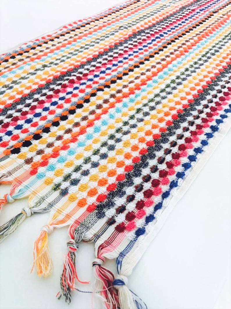 May include: A colorful, striped towel with a textured, dotted pattern. The towel features a variety of colors, including orange, blue, red, and white. The ends of the towel have decorative tassels.