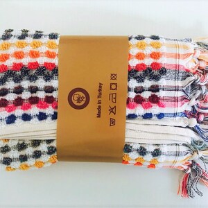 May include: Folded Turkish towel with a colorful, textured design. The towel features a pattern of small, raised dots in various colors, including orange, red, blue, and white. The towel is wrapped with a brown paper band that reads "Made in Turkey."