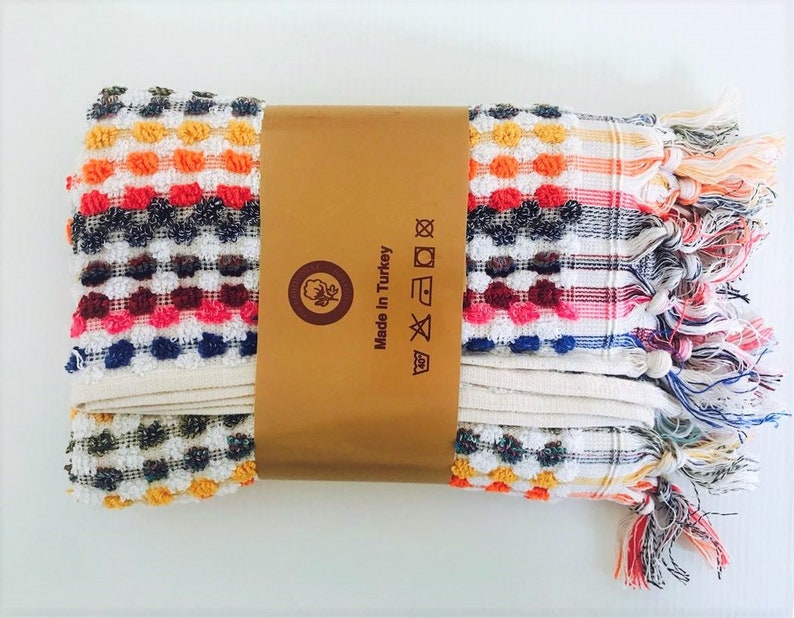May include: Folded Turkish towel with a colorful, textured design. The towel features a pattern of small, raised dots in various colors, including orange, red, blue, and white. The towel is wrapped with a brown paper band that reads "Made in Turkey."