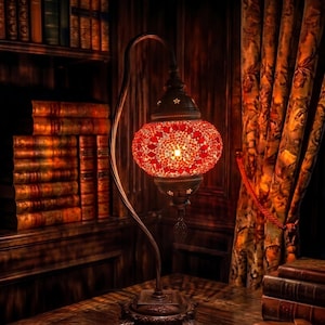 Puede incluir: Una lámpara de mesa decorativa con una pantalla de cristal de mosaico rojo, que emite un brillo cálido. La lámpara está sobre una mesa de madera oscura con libros y gafas, creando un ambiente acogedor de rincón de lectura. Estanterías y cortinas estampadas en el fondo.