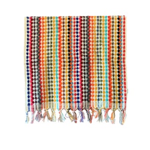 May include: A colorful striped towel with a textured dot pattern. The towel features vertical stripes in various colors, including red, orange, yellow, blue, and black. The bottom edge has decorative tassels.