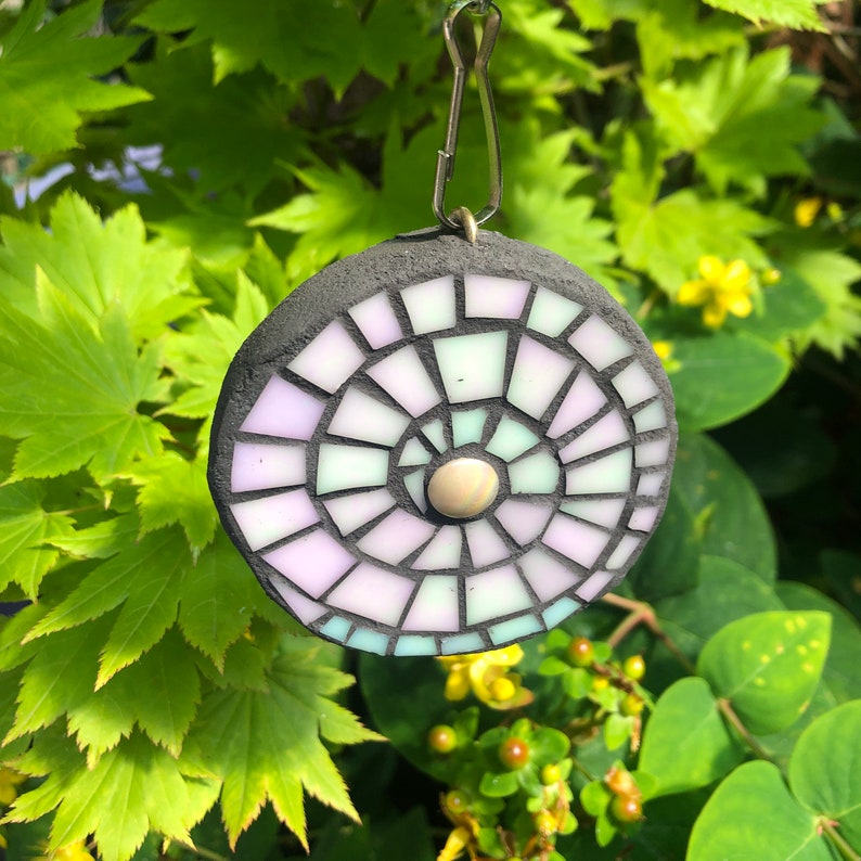 K&ouml;nnte beinhalten: Eine kreisrunde Mosaikfliese mit einem Spiralmuster in Violett-, Gr&uuml;n- und Wei&szlig;t&ouml;nen. Die Fliese ist an einem Metallh&auml;nger befestigt.