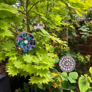 K&ouml;nnte beinhalten: Zwei runde Mosaik-Sonnenf&auml;nger, die an silbernen Ketten in einem Garten h&auml;ngen. Die Sonnenf&auml;nger bestehen aus schillernden Glasfliesen und haben eine wei&szlig;e Mitte. Die Sonnenf&auml;nger h&auml;ngen an einem Ast.