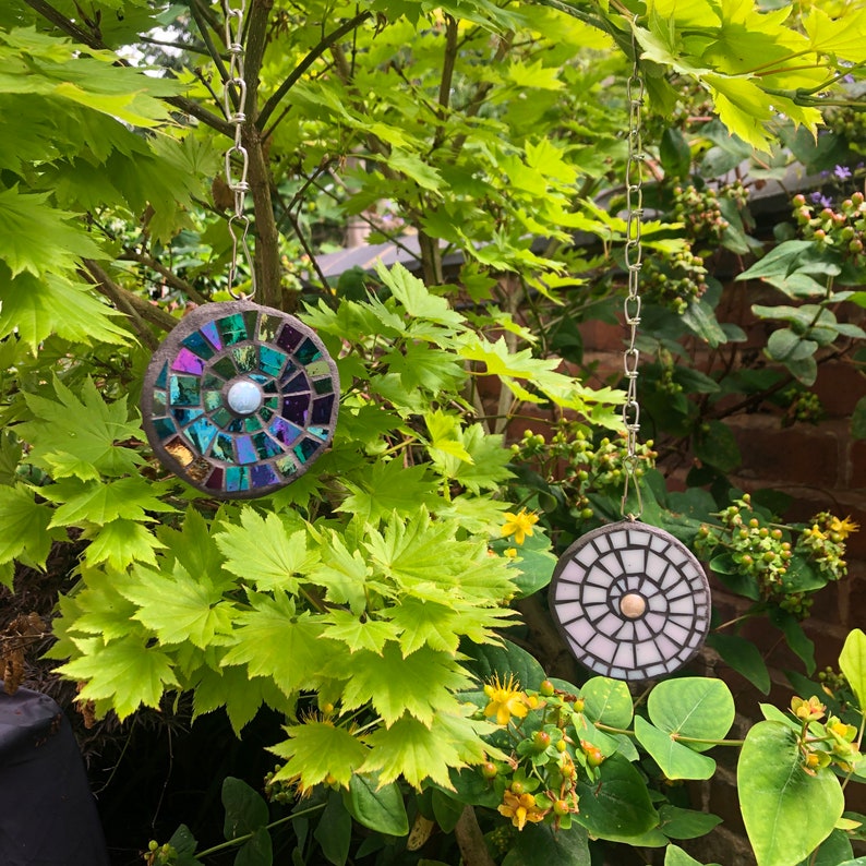 K&ouml;nnte beinhalten: Zwei runde Mosaik-Sonnenf&auml;nger, die an silbernen Ketten in einem Garten h&auml;ngen. Die Sonnenf&auml;nger bestehen aus schillernden Glasfliesen und haben eine wei&szlig;e Mitte. Die Sonnenf&auml;nger h&auml;ngen an einem Ast.