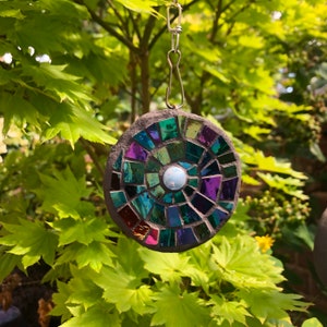 K&ouml;nnte beinhalten: Ein runder Mosaik-Sonnenf&auml;nger mit einem Spiralmuster in Gr&uuml;n-, Lila- und Blaut&ouml;nen. Der Sonnenf&auml;nger besteht aus kleinen Glasfliesen und hat eine wei&szlig;e, schillernde Mitte.