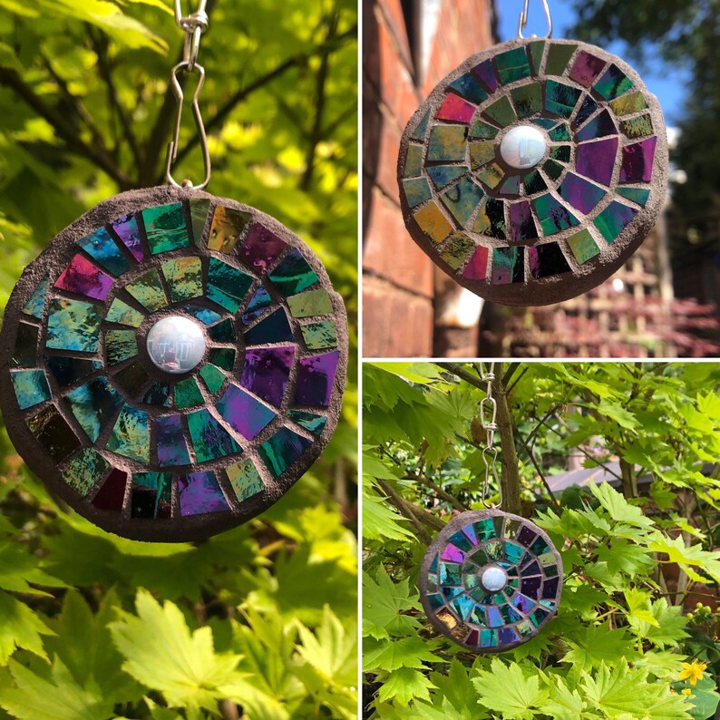 K&ouml;nnte beinhalten: Ein runder, schillernder Mosaik-Sonnenf&auml;nger mit einem Spiralmuster in Gr&uuml;n-, Lila- und Rott&ouml;nen. Der Sonnenf&auml;nger h&auml;ngt an einer silbernen Kette und hat eine kleine, reflektierende Scheibe in der Mitte.