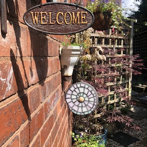 K&ouml;nnte beinhalten: Ein Holzschild mit dem Wort "WELCOME" in goldenen Buchstaben h&auml;ngt an einer Backsteinwand. Ein Mosaik-Sonnenf&auml;nger mit einem wei&szlig;en, rosa und blauen Design h&auml;ngt an einer Kette unter dem Schild.