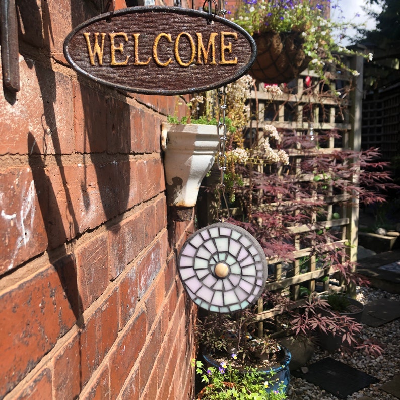 K&ouml;nnte beinhalten: Ein Holzschild mit dem Wort "WELCOME" in goldenen Buchstaben h&auml;ngt an einer Backsteinwand. Ein Mosaik-Sonnenf&auml;nger mit einem wei&szlig;en, rosa und blauen Design h&auml;ngt an einer Kette unter dem Schild.