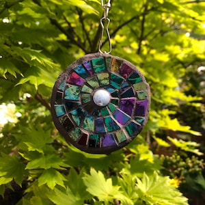 K&ouml;nnte beinhalten: Ein runder, schillernder Mosaik-Sonnenf&auml;nger mit dunkelgrauem Hintergrund und einem silbernen, reflektierenden Zentrum. Der Sonnenf&auml;nger h&auml;ngt an einer silbernen Kette.