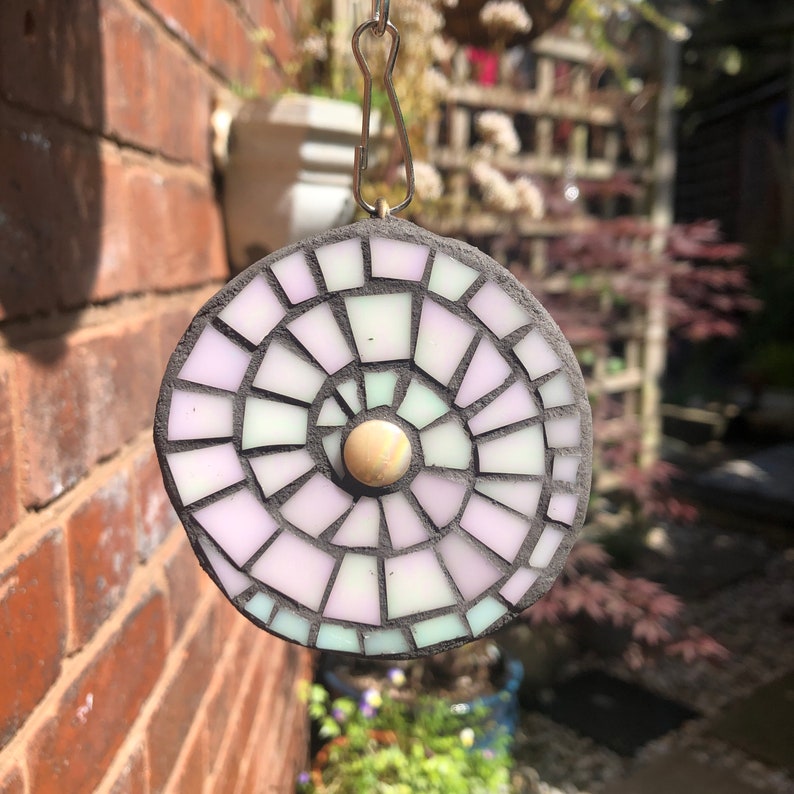 K&ouml;nnte beinhalten: Ein runder Mosaik-Sonnenf&auml;nger mit einem Spiraldesign in Rosa-, Wei&szlig;- und Gr&uuml;nt&ouml;nen. Der Sonnenf&auml;nger hat ein kleines, rundes, schillerndes Mittelst&uuml;ck.