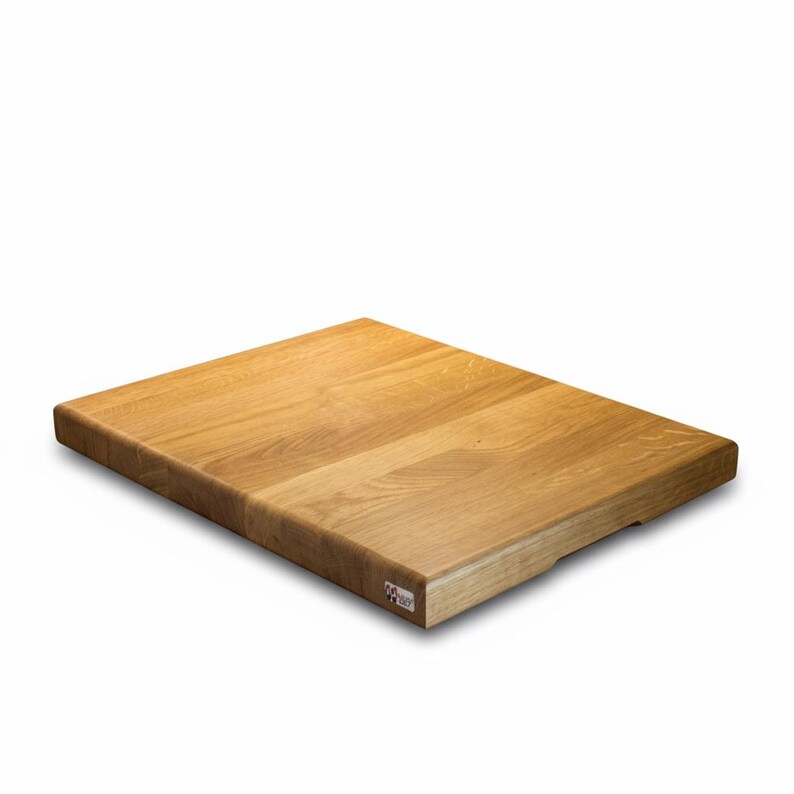 May include: A rectangular wooden cutting board with a light brown finish. The board has a smooth surface and a small, rectangular label in the lower right corner. The wood grain is visible, and the edges are slightly rounded.