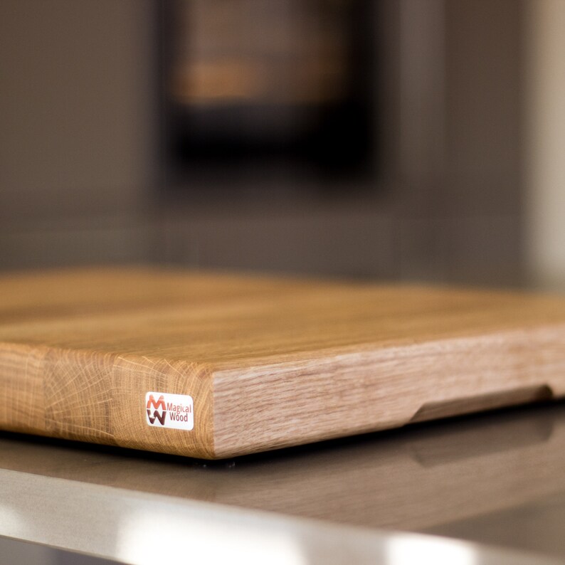 May include: A rectangular wooden cutting board with a light brown finish. The board has a beveled edge and a small cutout on one side. A sticker with the text "Magical Wood" is on the corner.