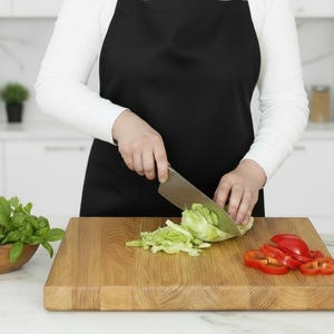 May include: A person wearing a black apron is cutting lettuce on a wooden cutting board. Sliced red bell peppers and a wooden bowl of green herbs are also on the board. The kitchen has white cabinets and appliances.