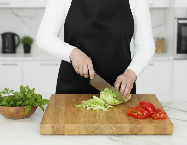 May include: A person wearing a black apron is cutting lettuce on a wooden cutting board. Sliced red bell peppers and a wooden bowl of green herbs are also on the board. The kitchen has white cabinets and appliances.
