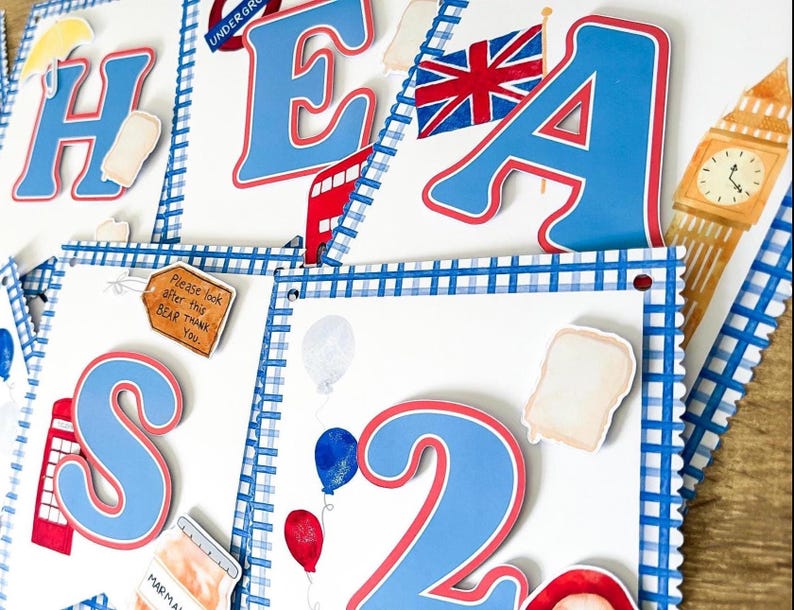 May include: A collection of decorative paper cutouts with a British theme. The cutouts include the letters "H E A S 2", a Union Jack flag, a red double-decker bus, Big Ben, and various food and balloon illustrations. The letters are blue with red trim.